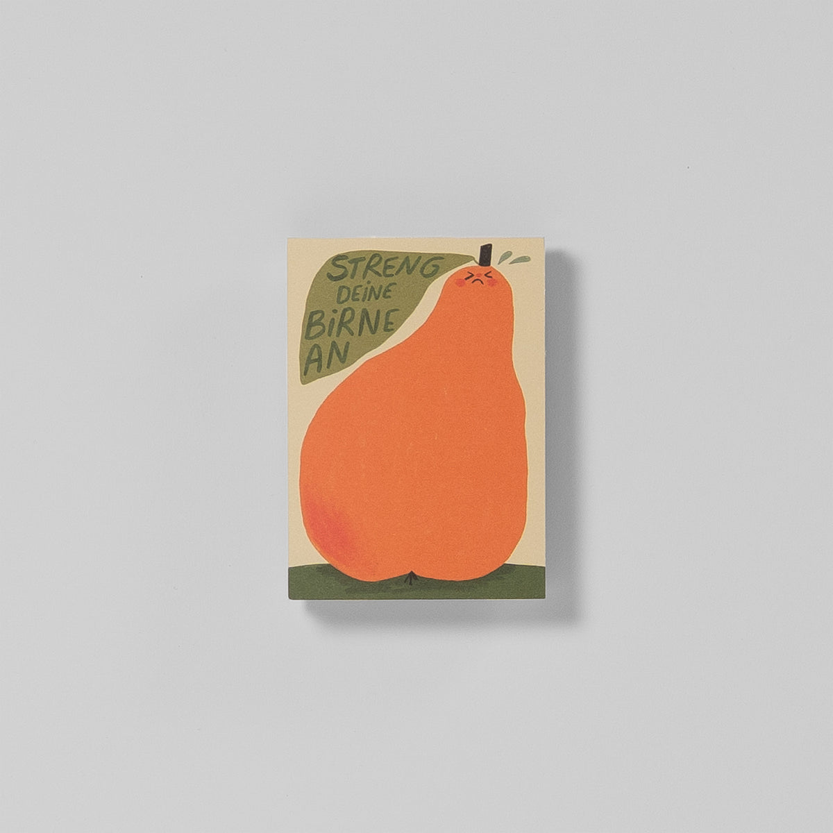 Notizblock A7 „Streng deine Birne an“ von Illustratorin Slinga – cremefarbener Hintergrund, orangene Birne mit grimmigem Gesicht auf grüner Wiese