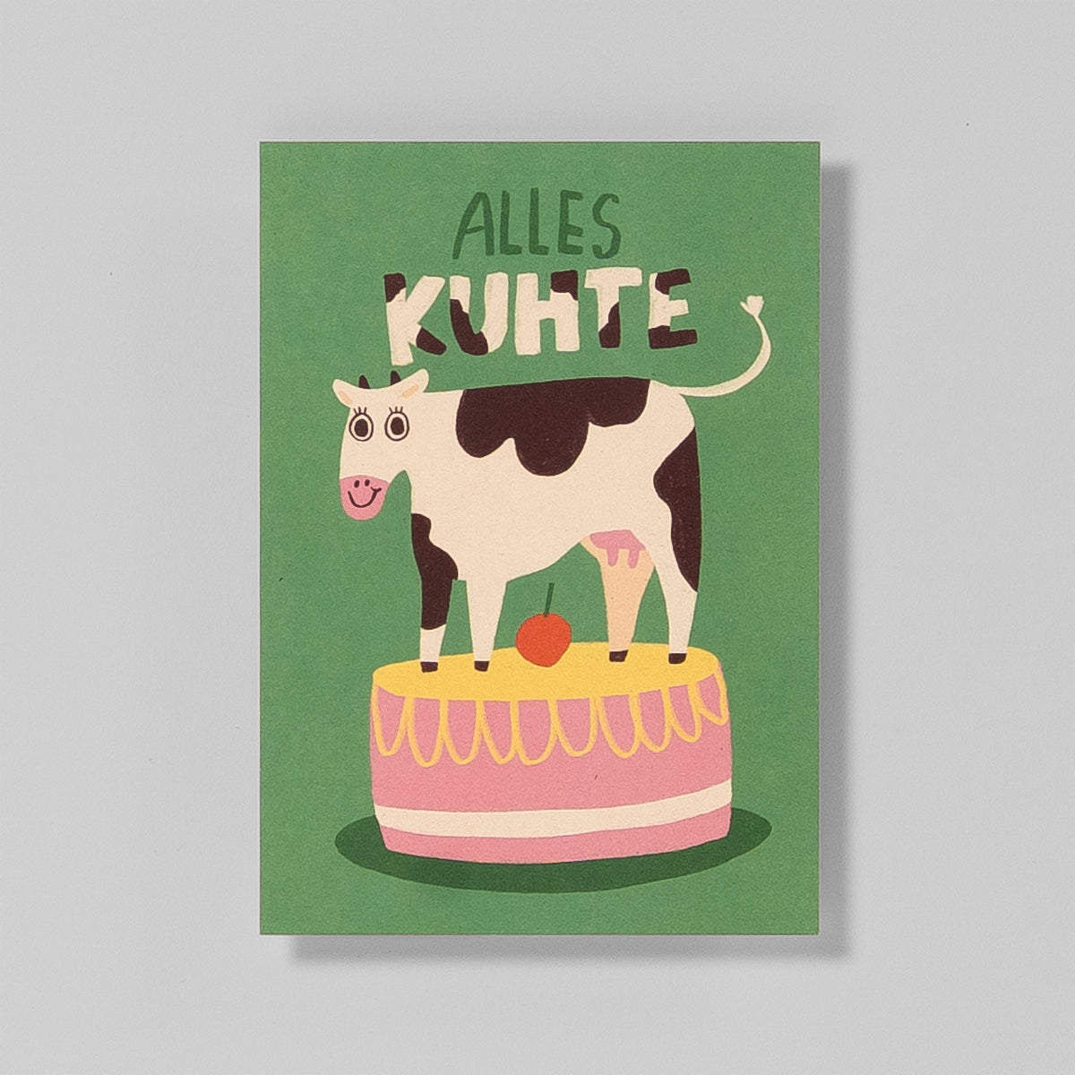Postkarte A6 „Alles Kuhte“ von Illustratorin Slinga – grüner Hintergrund, schwarzweiße Kuh steht auf Geburtstagstorte und freut sich