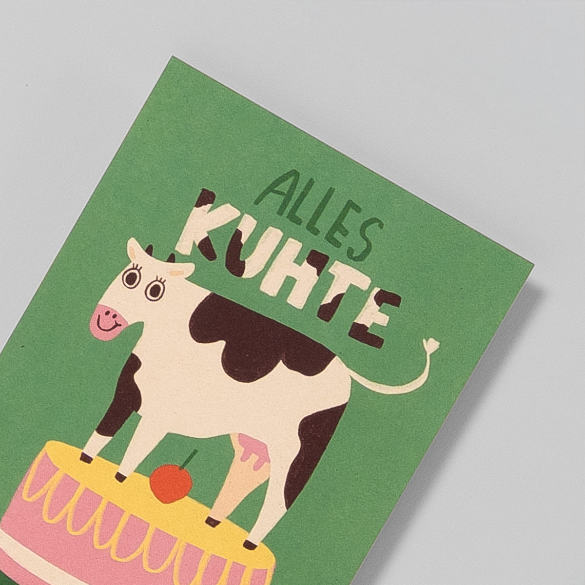 Postkarte A6 „Alles Kuhte“ von Illustratorin Slinga – grüner Hintergrund, schwarzweiße Kuh steht auf Geburtstagstorte, Nahaufnahme Kuh und Überschrift